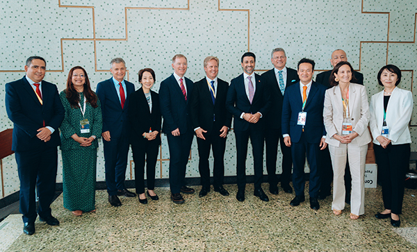 Fourteen people wearing business attire and conference ID badges stand in a line indoors for a formal group photograph, with a patterned wall behind them.