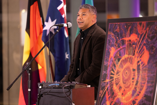 Person speaking at a lectern during a NAIDOC Week event, with Aboriginal and Torres Strait Islander flags visible in the background and artwork displayed beside the stage.