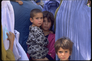 A girl holds a younger boy, standing amidst other children and women dressed in burqa.