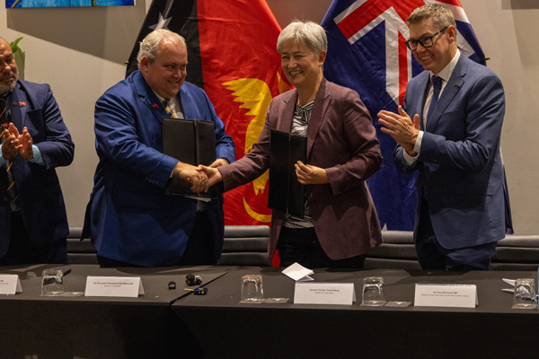 Two people shake hands while holding document folders, with Papua New Guinea and Australian flags behind them as other attendees applaud.