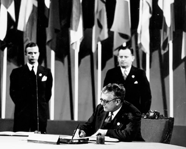 Black-and-white photograph shows a suited official signing documents at a conference table, with two attendants standing behind him. Rows of national flags create an international diplomatic setting.