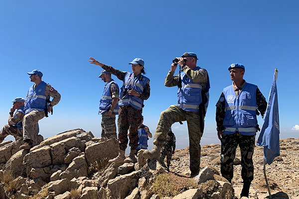 Photograph shows uniformed field personnel in blue vests surveying a rocky, arid landscape under clear skies. Group includes members using binoculars and gesturing toward distant terrain, with a blue flag marking their position.