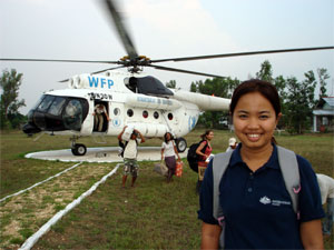 AusAID officer standing in the foreground smiling and behind her is a World Food Programme helicopter transporting people