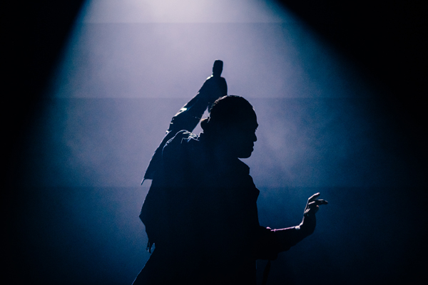 Photograph of a person performing on stage, captured in silhouette against a blue-lit background. The figure's raised arm and poised hand suggest a dramatic or expressive moment during a dance or theatrical performance.