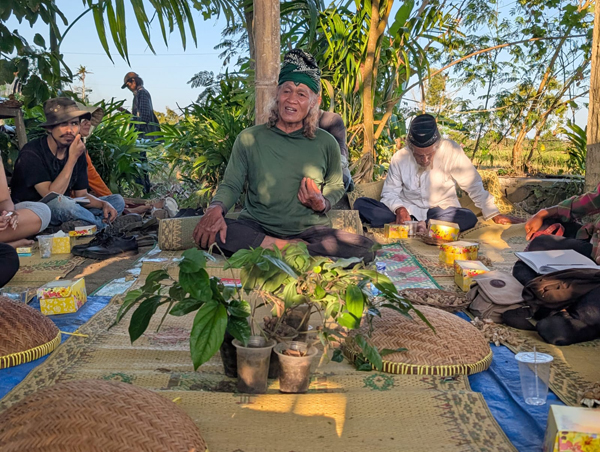 Photograph of a group of people sitting on mats outdoors, engaging in a communal gathering or discussion surrounded by plants and trees. Central figure wears a green shirt and traditional Indonesian headwear, with various items like food containers and potted plants arranged around participants, highlighting a cultural or social event in a natural setting.