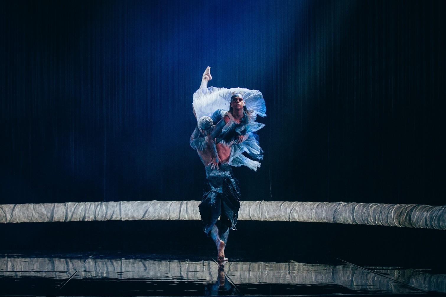 Photograph of two Indigenous dancers performing on stage with dark blue lighting and a reflective floor. The male dancer lifts the female dancer, who wears a flowing white costume and points upward, highlighting grace and strength in a dramatic moment.