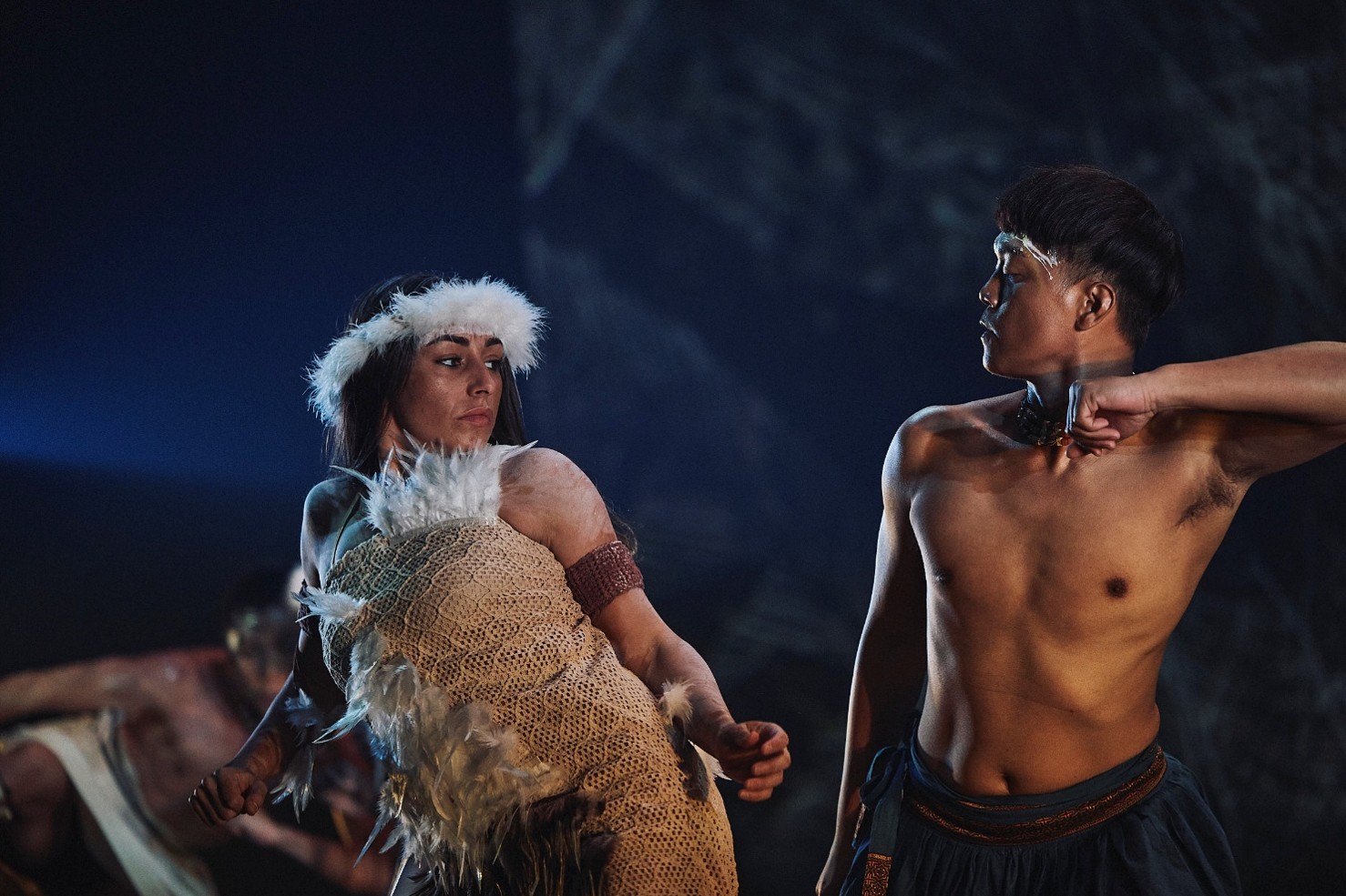 Photograph of two dancers performing a traditional or cultural dance on stage with dark background. The female dancer wears a fur headpiece and woven garment, while the male dancer is shirtless with dark pants, both striking dynamic poses.