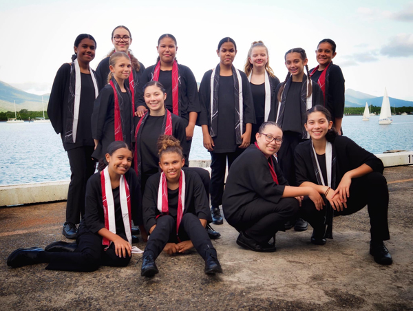 Photograph of a group of twelve young Indigenous girls posing outdoors near a body of water with sailboats in the background. They wear coordinated black outfits with red and white scarves, suggesting participation in a team or performance group.