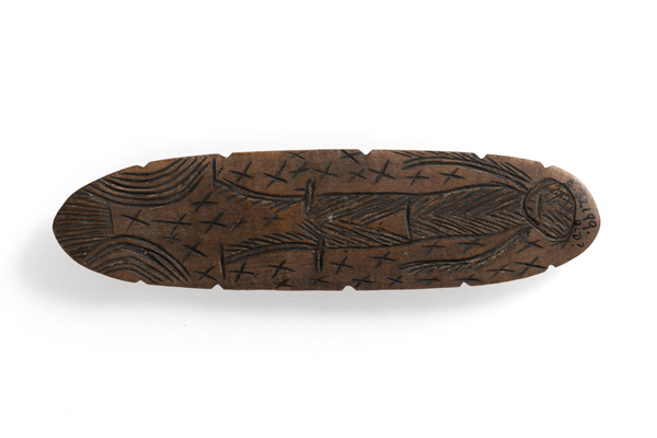 Photograph of a carved wooden artifact shaped like an elongated oval with a human figure etched in the center. The carving features cross patterns surrounding the figure and linear designs on both ends, highlighting traditional craftsmanship.