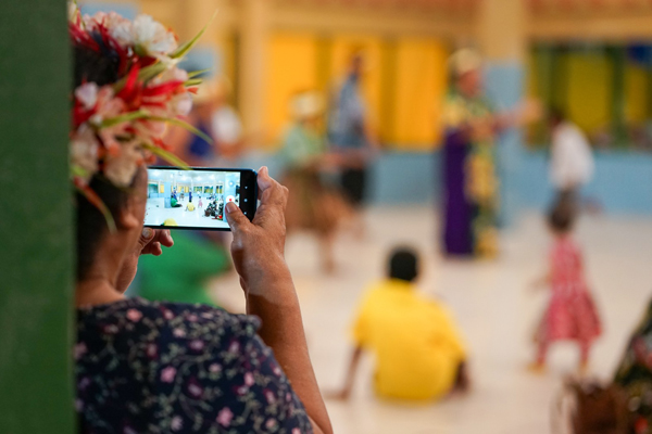 Photograph showing a person with a floral headpiece capturing a cultural dance performance on a smartphone. The scene includes dancers in traditional attire and children seated or standing in a brightly lit indoor space with blue and yellow walls.