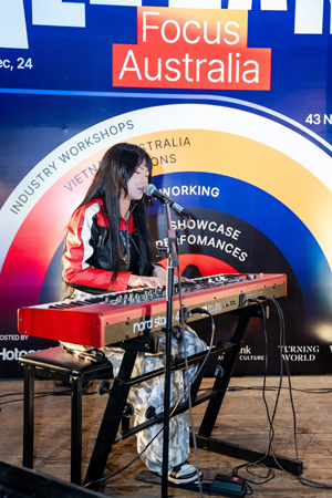Photograph of a female Vietnamese musician playing a red Nord Stage keyboard and singing into a microphone at an event with a colorful circular backdrop. Backdrop features labeled concentric rings in white, yellow, blue, and black with text such as "Industry Workshops," "Australia," "Networking," and "Showcase Performances," highlighting event focus on Australian music and cultural exchange.