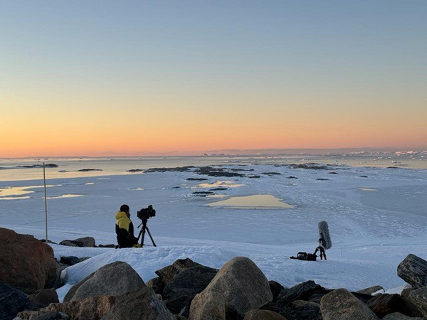 Photograph of a snowy coastal landscape at sunset with a person in a yellow jacket operating a camera on a tripod and audio equipment set up nearby. Large rocks in foreground contrast with icy water and scattered ice patches extending toward horizon under a clear sky with warm orange and blue hues.