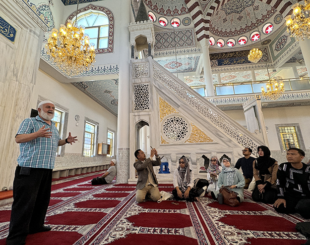 Group of people seated on a carpet listening to a religious leader speaking in a mosque.