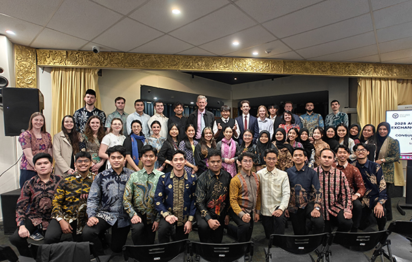 Large group photo of program delegates standing and seated in rows inside a formal event space with a stage backdrop and decorative detailing.