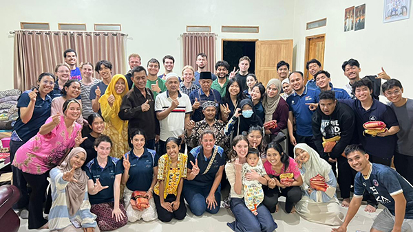 Program delegates and host community members gathered indoors at a family home, arranged in a large group with some seated and others standing, creating a welcoming group portrait.