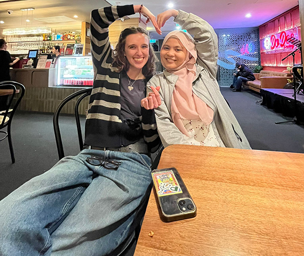 Two exchange program delegates seated at a café table, forming a heart shape with their arms, with the café interior visible in the background.