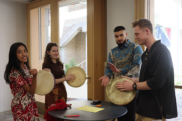 Program delegates standing around a small table indoors while playing traditional Indonesian hand drums during a cultural activity session.