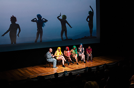 Six performers seated on stage under lights.
