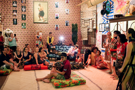Man seated on cushion playing guitar in middle of room, surrounded by others listening, watching.
