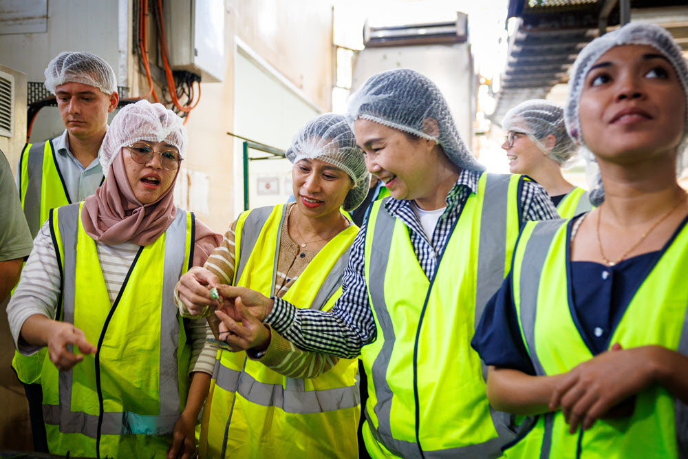 Photograph shows diverse workers in fluorescent safety vests and hairnets touring an industrial facility. A woman gestures toward a small object while colleagues observe, highlighting workplace training, inspection, or manufacturing processes.