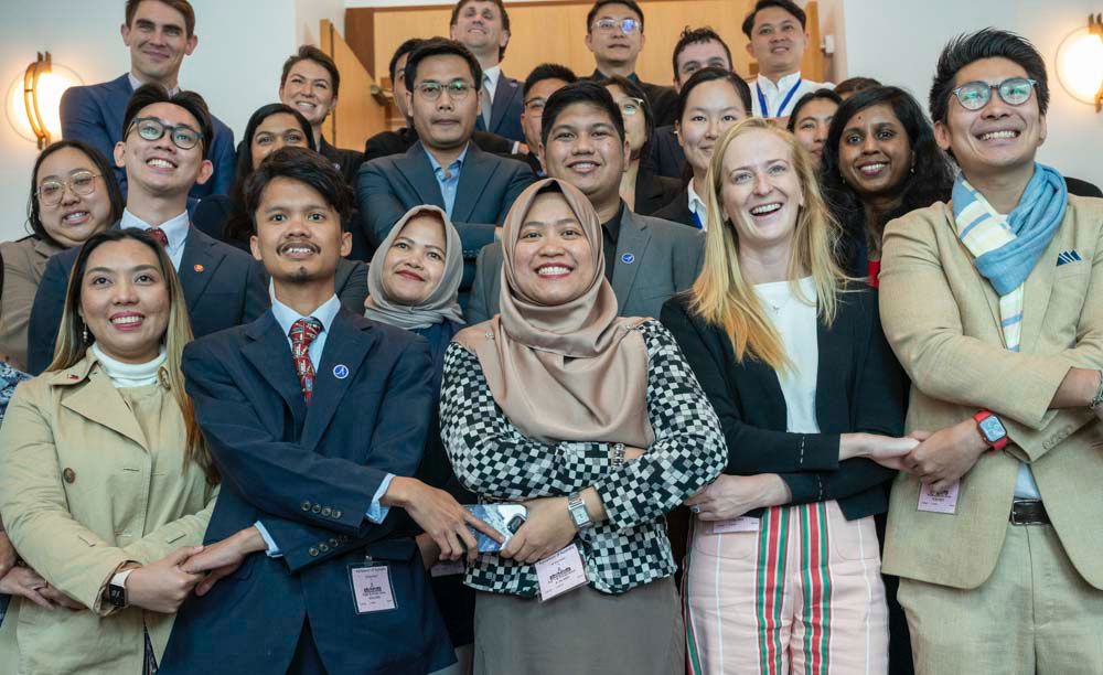 Group photograph shows a diverse group of smiling professionals gathered indoors, many wearing business attire, scarves, and conference badges. Front row features several women and men, with additional colleagues arranged behind them, suggesting a workplace, conference, or team gathering.