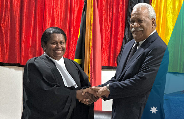 Two individuals shaking hands in front of red curtains and multiple flags; one wearing formal judicial attire and the other in a dark suit, during a ceremonial or official event.