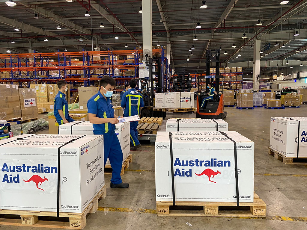 Men wearing masks are inspecting a delivery of vaccines stored in a white coloured temperature-controlled box with the stickers ‘Australian Aid’