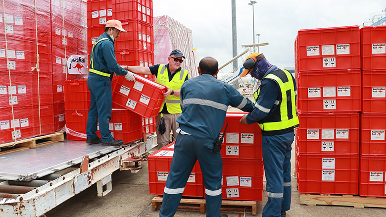 Men wearing high-visibility clothing are unloading humanitarian supplies. They are red boxes, known as ‘Kitchen Kits’, and have ‘Australian Aid’ stickers on them.