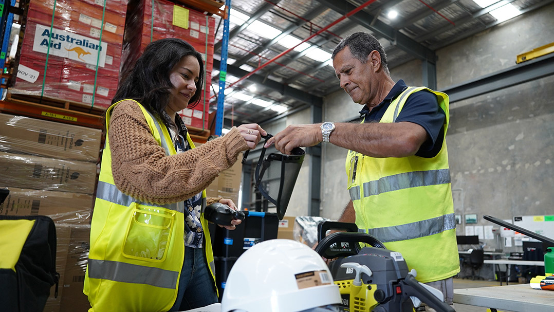 A woman on the left and a man on the right, both wearing high-visibility vests, hold a face protection visor, which is attached on helmets for safety.
