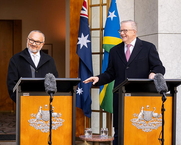 Two adults stand at podiums with the Australian coat of arms, speaking at an outdoor press conference. Australian and Solomon Islands flags hang behind them, with microphones on stands in front.