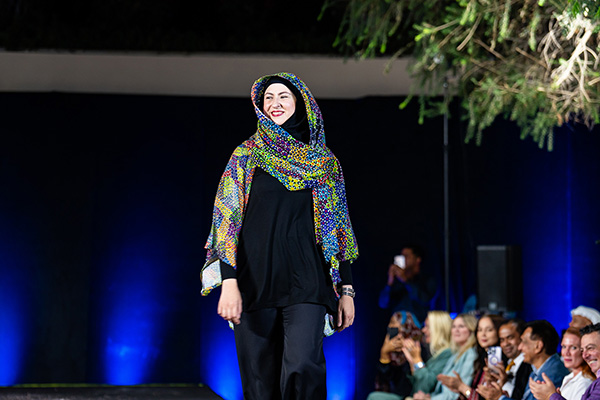 Person walking on a runway wearing a black outfit paired with a colorful patterned scarf, with an audience seated in the background.
