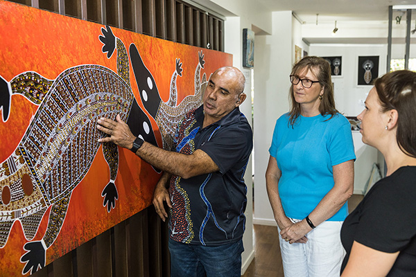 Person pointing to details on a large Aboriginal artwork featuring two lizards with intricate dot patterns, while others observe.