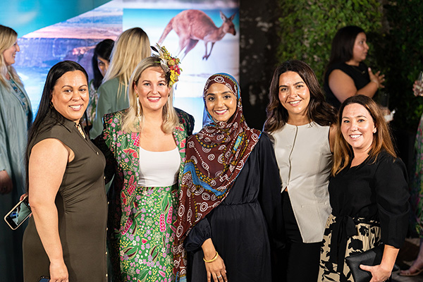 A group of people standing together at an event, dressed in a mix of formal and cultural attire. One person wears a green patterned outfit with a floral headpiece, another wears a dark outfit with a patterned scarf. The background features a large banner with an image of a kangaroo and a scenic landscape, along with greenery on one side.