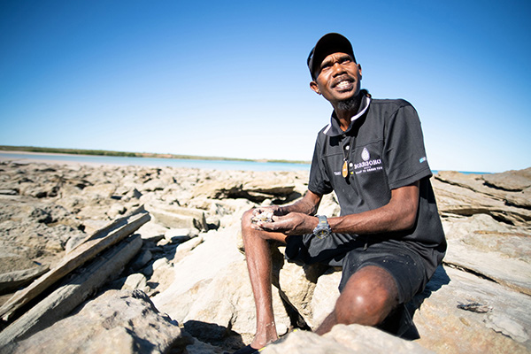 Person sitting on rocky terrain near a coastal area under a clear blue sky, holding small shells or stones in their hands. The background shows calm water and a distant shoreline.