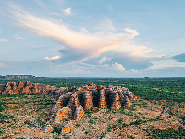 Aerial view of the Bungle Bungle Range in Purnululu National Park, Western Australia. The scene shows striking beehive-shaped sandstone formations with orange and gray banding, surrounded by green vegetation. The sky above is expansive with soft clouds illuminated by sunlight, creating a dramatic backdrop.