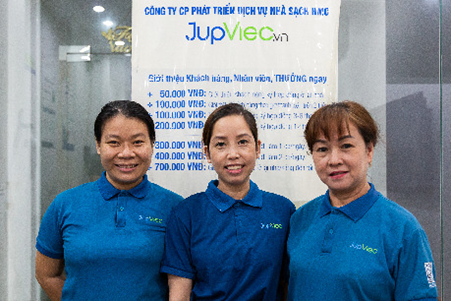 Three Vietnamese women in blue polos smiling at camera in front of a JupViec poster with Vietnamese writing.