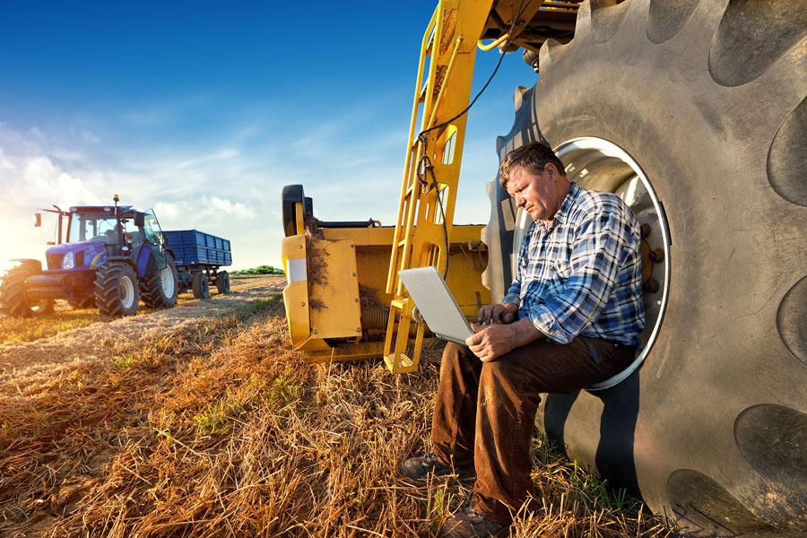 Photo of a man sitting in a large tractor tire with a laptop on his lap in a large farm field. Tractor plowing in the background. 