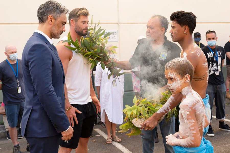Photo of Movie director Taika Waititi and Actor Chris Hemsworth accepting a smoking leaves blessing from an Indigenous Australian during an Indigenous land acknowledgment ceremony.