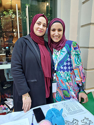 Two individuals stand side by side in front of a glass window. One wears a dark jacket, the other a colourful patterned shirt. Both wear maroon headscarves. A table nearby holds animal-themed colouring pages and pens.