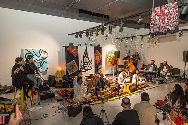 Live musical performance in an indoor venue decorated with art and fabric hangings. Musicians seated on the floor play traditional instruments like harmoniums and drums, while an audience watches from chairs and the floor. Spotlights and decorative ceiling lights illuminate the space.