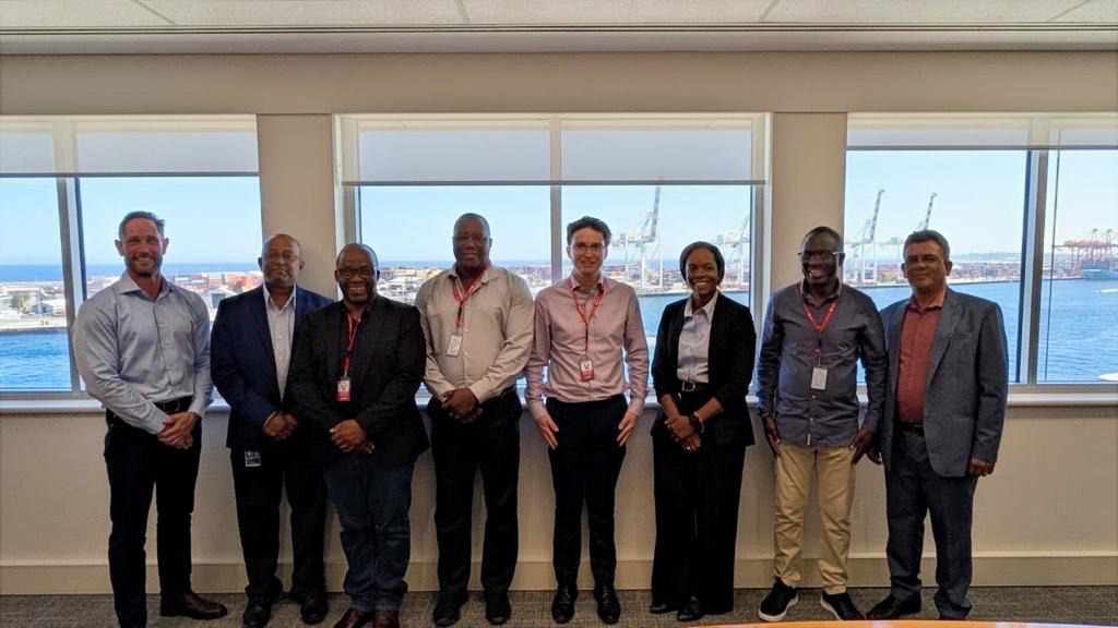 Eight individuals in business attire stand side by side indoors for a photo. They are standing in front of windows with a view of Fremantle harbour.