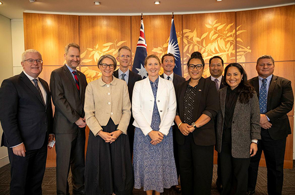 A group of officials in formal attire smiling in front of the Australian and Marshall Islands flags, with a textured wooden wall as the backdrop.