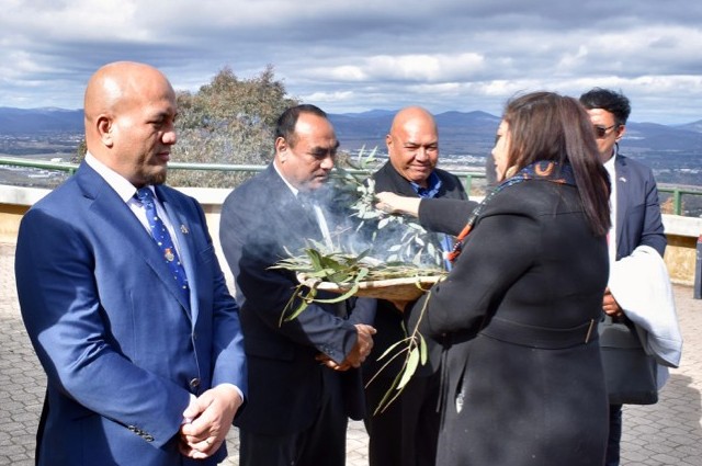 Four individuals in suits at Mount Ainslie in Canberra during a Welcome to Country ceremony, with one engaging in the tradition by smelling smoke from burning eucalyptus leaves.