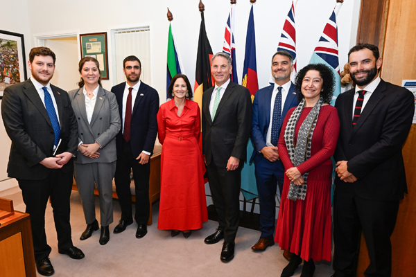 Seven people in business attire, alongside one Australian Minister, standing in front of several national flags pose for a formal group photo.