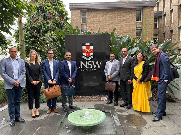 A group of eight people standing in front of a UNSW Sydney sign, with plants and buildings in the background.