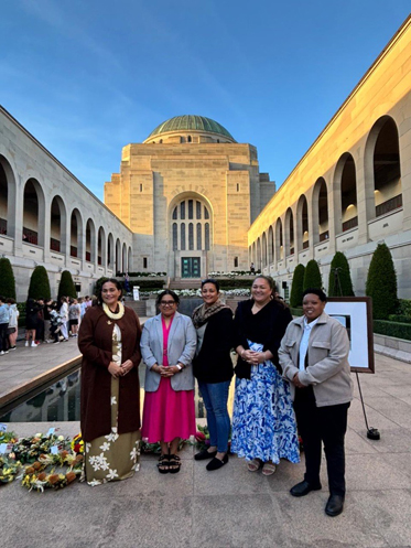 Five females from the Pacific stand side by side in an open-air courtyard. The group is positioned in front of the reflective pool and a line of commemorative wreaths.