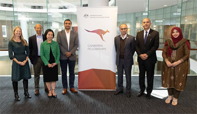 Seven people in business attire stand in a row, posing for a photo beside a Canberra Fellowships Program banner in a modern office foyer
