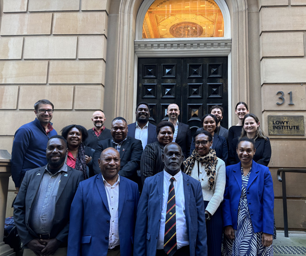  group of sixteen individuals in work attire stand in front of an office building, smiling towards the camera.