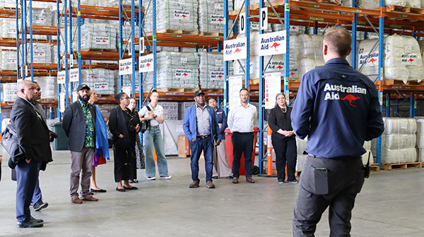 A group of people stand in a warehouse of Australian Aid humanitarian emergency relief supplies, listening to an officer giving a tour of the warehouse. 