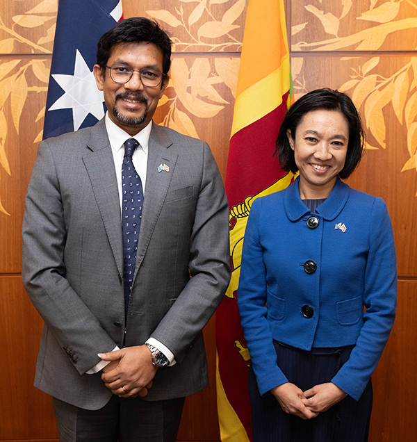 Two officials in formal attire smiling in front of the Australian and Sri Lanka flags, with a gumtree-leaf textured wooden wall as the backdrop.